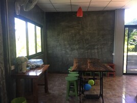 Una habitación con una gran mesa de madera, taburetes verdes, un ventilador y una lámpara colgante roja.