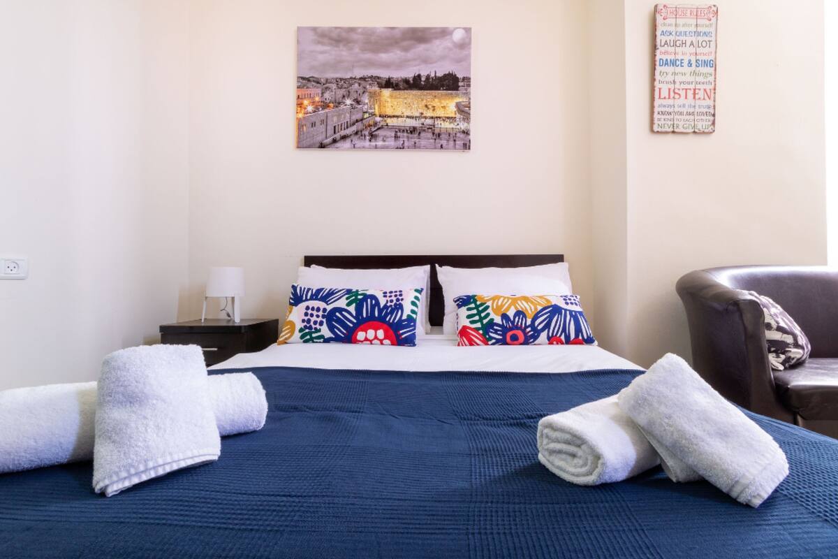 Une chambre d’hôtel avec un lit, deux oreillers, une table de chevet, une lampe, une chaise et un mur orné d’un tableau représentant un paysage urbain et d’une citation inspirante.