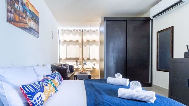 A hotel room with a bed, a desk, a chair, a wardrobe, and a painting on the wall. - NHE Machne Yehuda Apartments (Jerusalem)