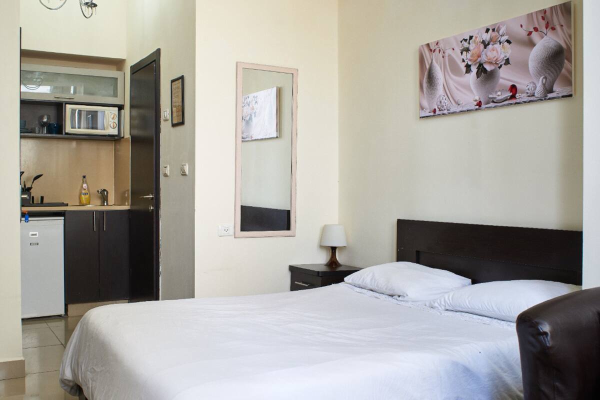 Une chambre d’hôtel comprenant un lit, une table de chevet, une lampe, un miroir fixé au mur, un tableau, une kitchenette équipée d’un four à micro-ondes et d’un réfrigérateur.