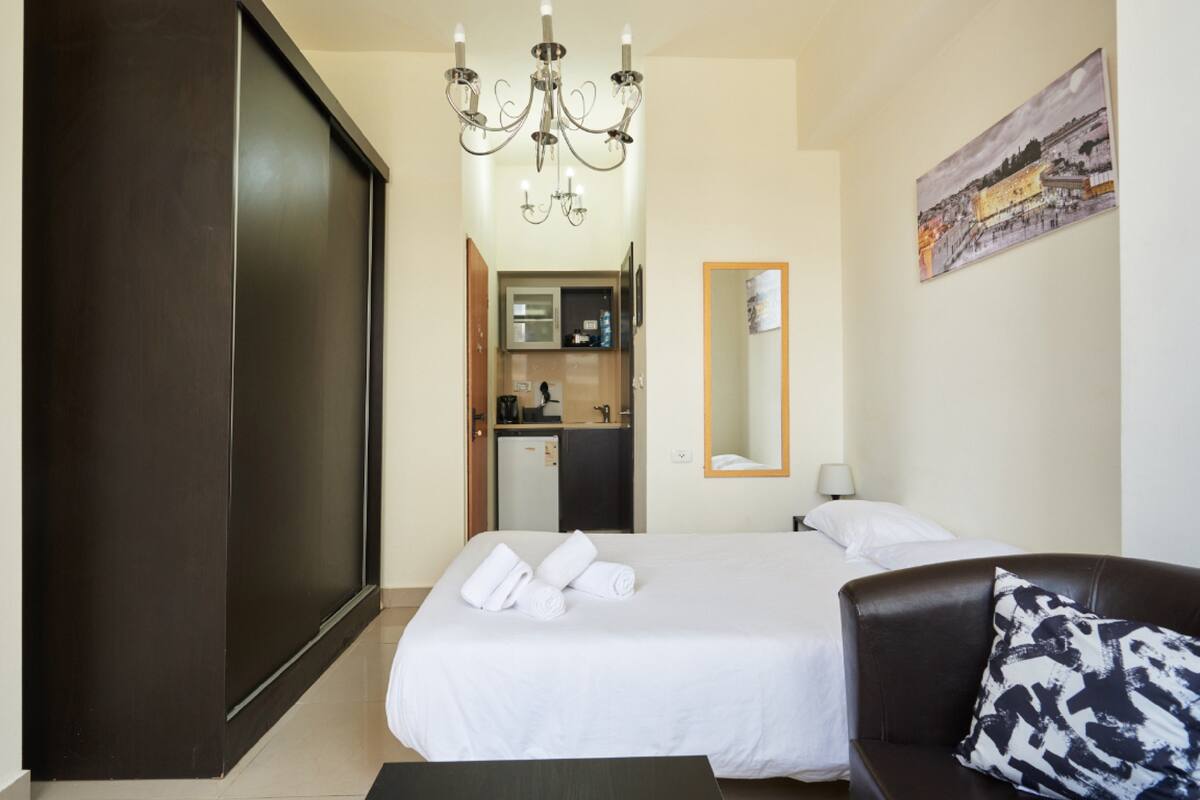 Une chambre d’hôtel avec un lit, une armoire en bois sombre, un lustre, un tableau et, au fond, une petite cuisine.