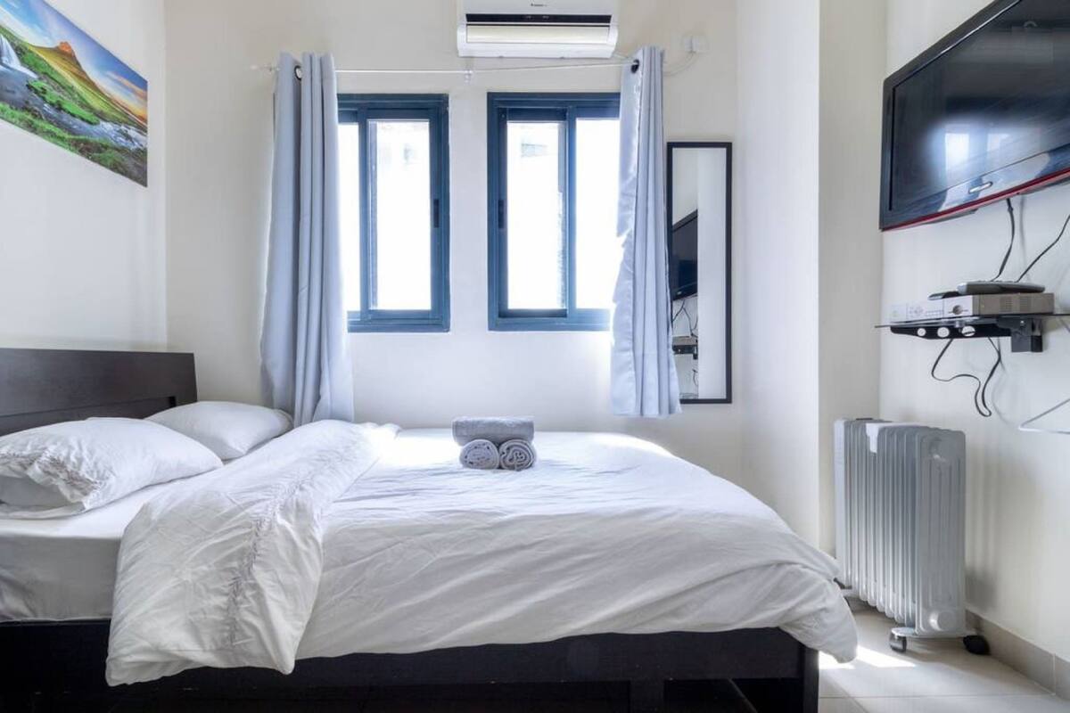 Une chambre à coucher avec un lit, deux fenêtres, un climatiseur, une télévision et un radiateur.