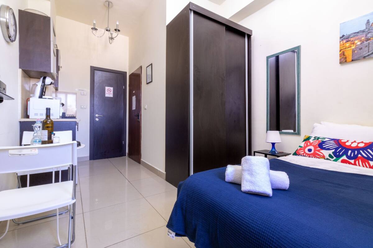 Une chambre d’hôtel avec un lit, un bureau, une chaise, une armoire, un miroir et une horloge au mur.