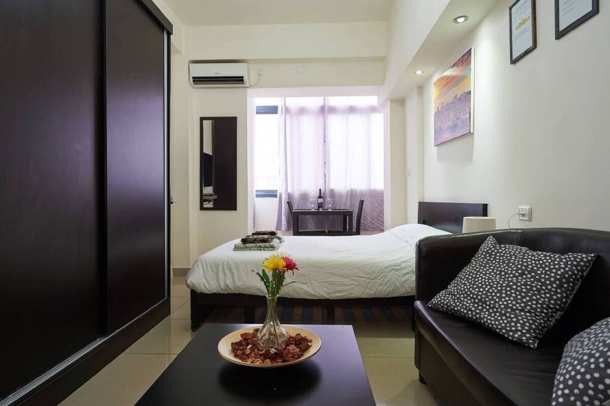 Une chambre d’hôtel avec un lit, un canapé, une table basse avec un vase et une fenêtre avec des rideaux.