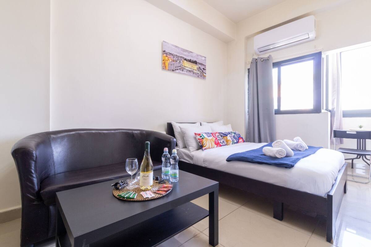 Une chambre d’hôtel avec un lit, un canapé, une petite table sur laquelle se trouvent une bouteille de vin, des verres et des bouteilles d’eau.