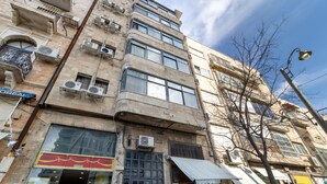 A multi-story building with air conditioning units, a storefront with a red awning, and a tree in front. - NHE Machne Yehuda Apartments (Jerusalem)