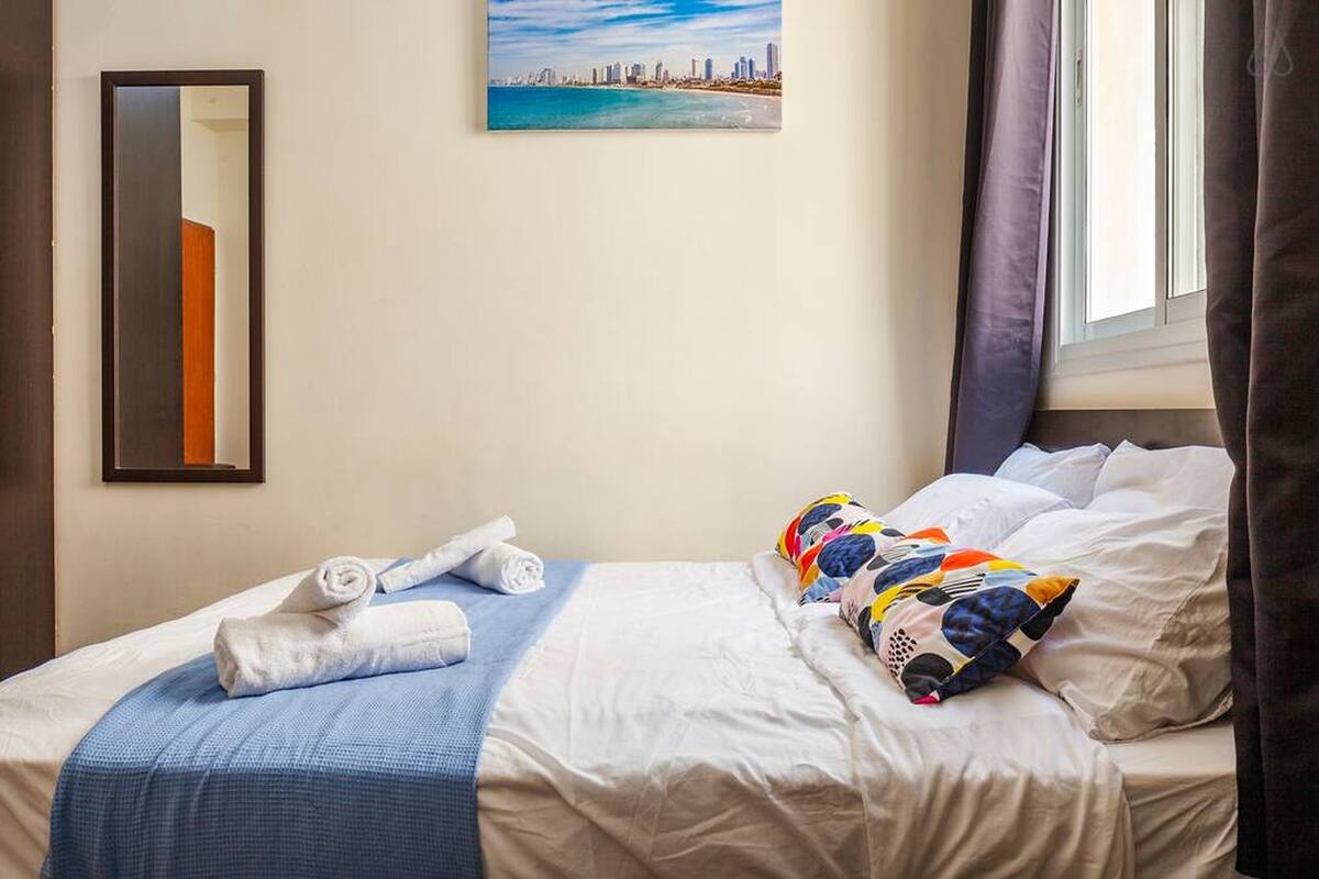 A hotel room with a bed, a window with curtains, a painting on the wall, and a mirror.