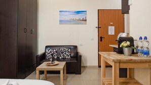A compact room with a black leather sofa, a wooden coffee table, a bed, a dark wardrobe, a wooden door, and a small table with a kettle and water bottles. - NHE Rothschild Apartments (Tel Aviv)