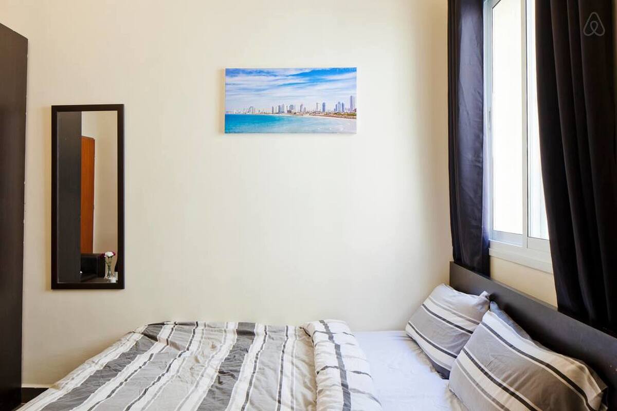 A hotel room with a bed, a cityscape painting, a mirror, and a window with curtains.