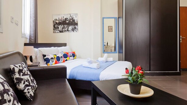 A hotel room with a bed, a sofa, a table with a plant, a wardrobe, and a framed photo on the wall. - NHE Rothschild Apartments (Tel Aviv)
