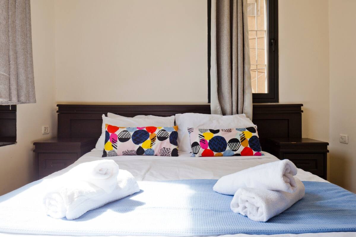 Un lit bien fait, avec des draps blancs et des coussins colorés, une couverture bleue et deux serviettes enroulées sur un tapis bleu.