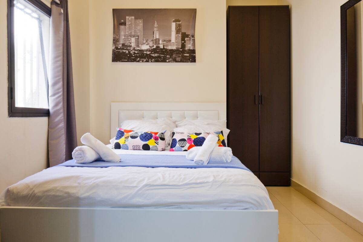 Une chambre d’hôtel avec un lit, un tableau représentant un paysage urbain accroché au mur, une armoire et une fenêtre avec des rideaux.
