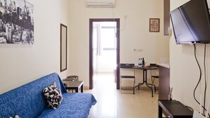 Une chambre d’hôtel avec un canapé bleu, une petite table et une chaise, un téléviseur et un bureau avec une chaise. Une porte donne sur un couloir lumineux.
