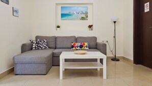 A living room with a grey sofa, a white coffee table, a painting of a beach scene, a floor lamp, and a door with a no-smoking sign. - NHE Rothschild Allenby Apartments (Tel Aviv)