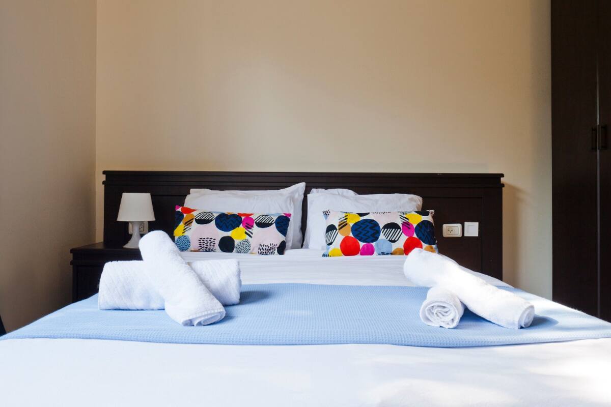Un lit bien fait, recouvert d’une couverture bleue, avec deux oreillers aux motifs colorés et deux serviettes blanches roulées.