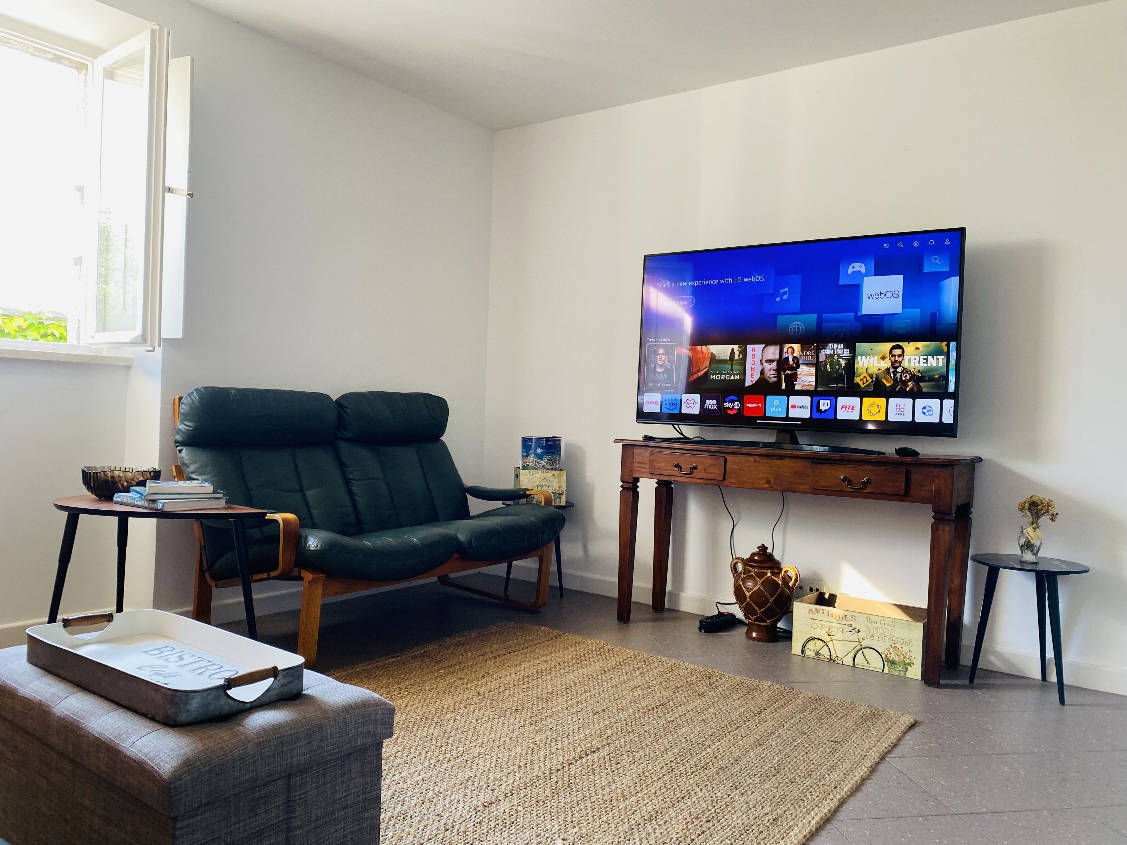 Una sala de estar con un televisor de pantalla plana grande montado sobre un soporte de madera, un sillón de cuero, una mesita auxiliar con libros y un jarrón decorativo en una mesita.
