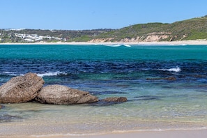 A coastal beach with clear turquoise water, rocky formations, and a distant shoreline with buildings.