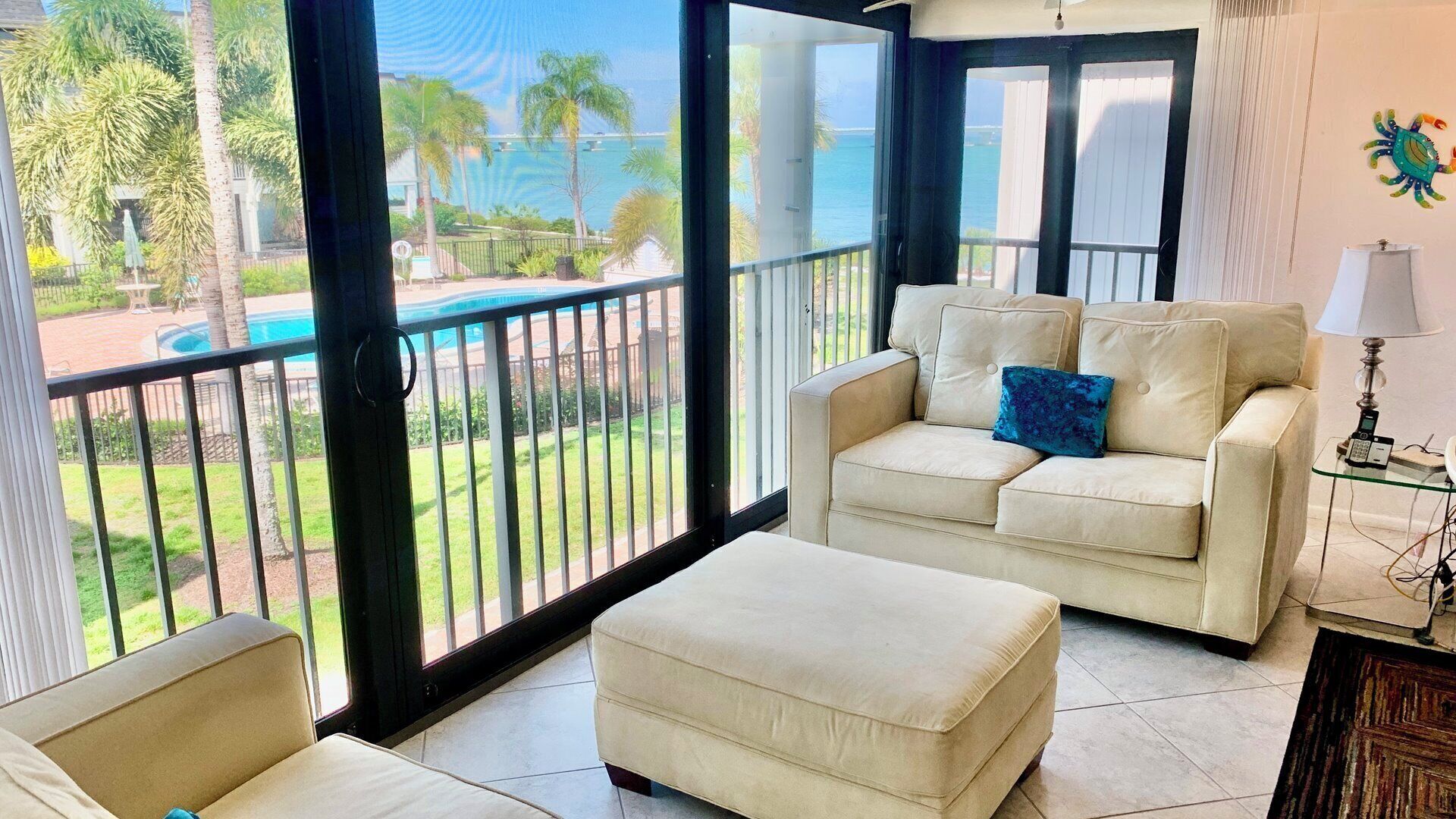 A room with a beige sofa, a matching ottoman, and a view of a pool and palm trees.
