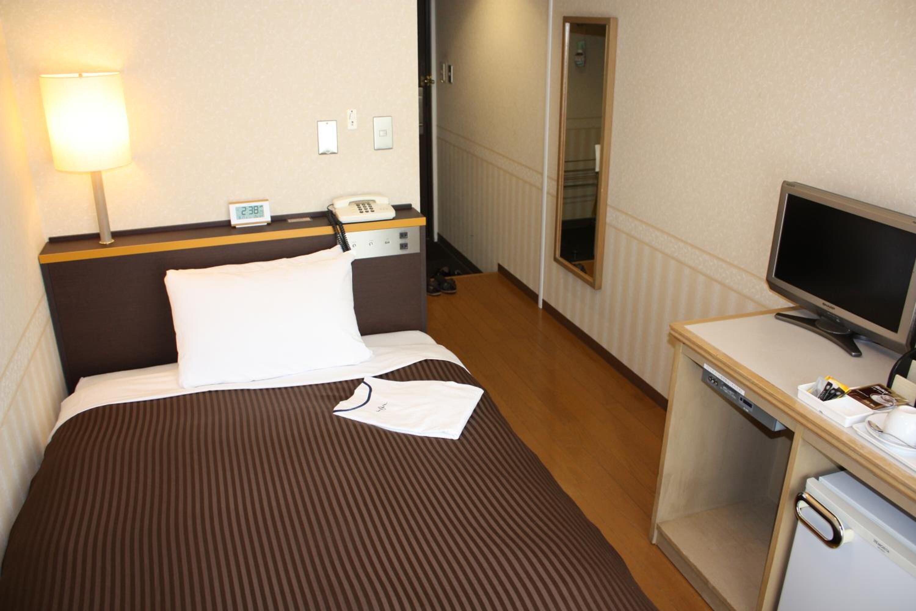A hotel room with a bed, a bedside lamp, a desk with a computer, and a mirror.