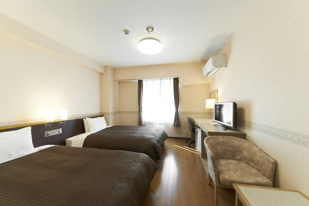 Habitación de hotel con dos camas, un escritorio con televisor, una silla y una ventana con cortinas.