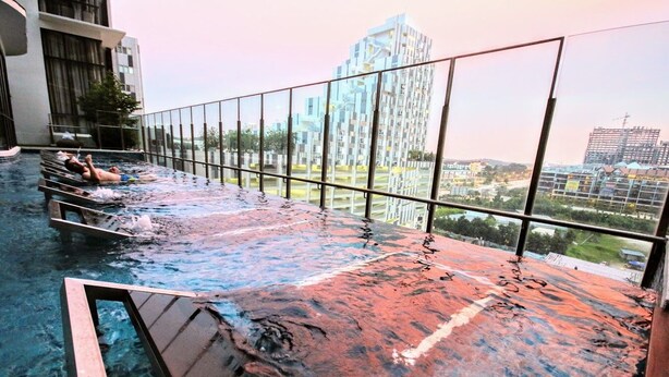 Una piscina infinita en la azotea con barandilla de vidrio, que ofrece vistas de edificios circundantes y una obra en construcción.