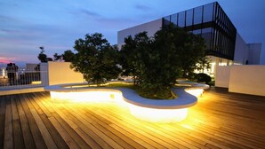 Una terraza en la azotea con cubierta de madera, un edificio moderno y una zona ajardinada con un árbol y asientos.