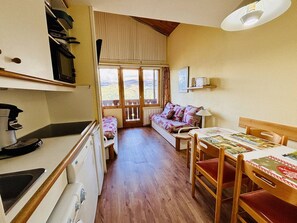 Ein gemütliches Zimmer mit Holzboden, einer Couch, einem Esstisch mit Stühlen, einer Küchenzeile mit Herd und einem großen Fenster mit Aussicht.