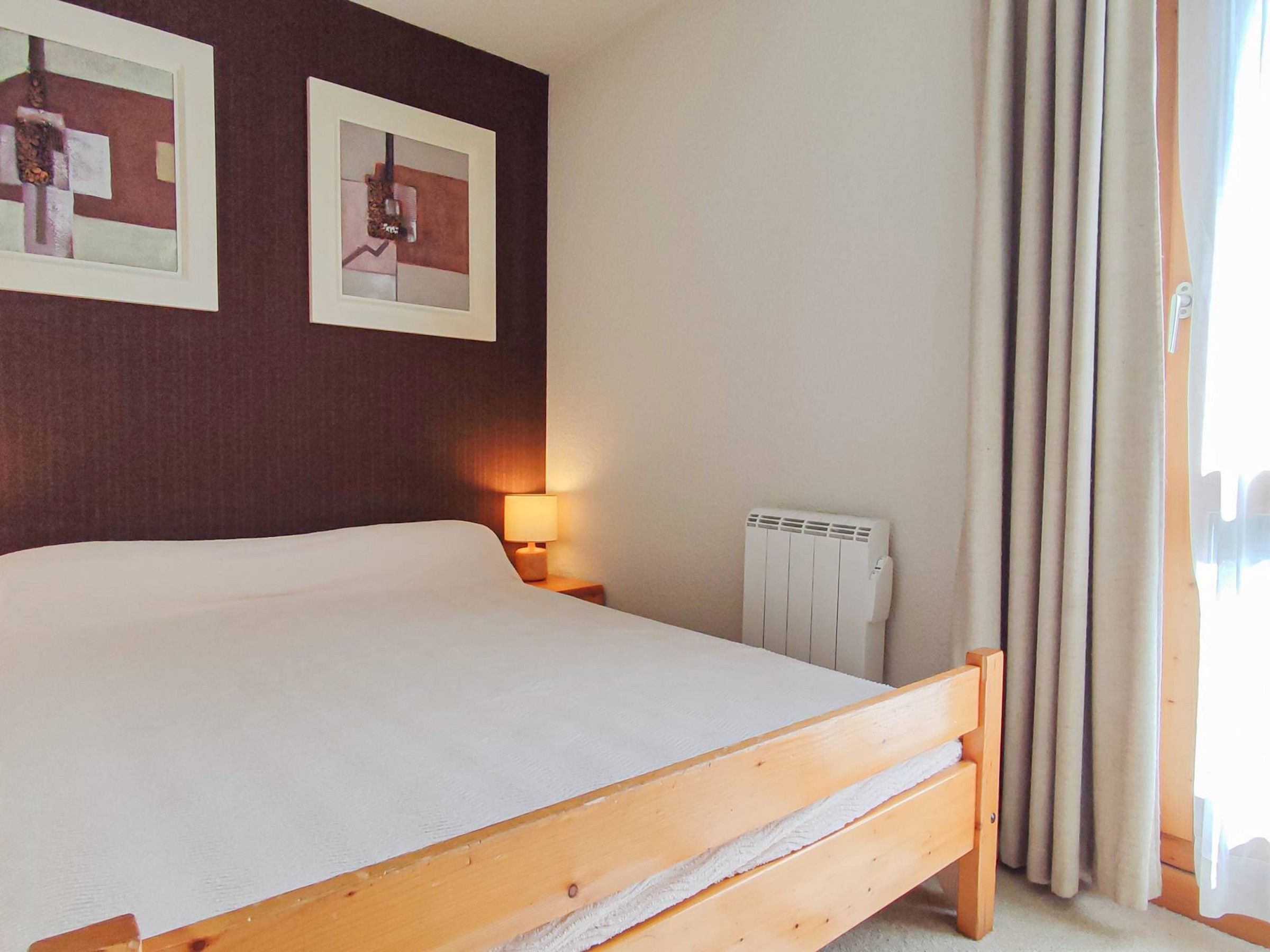 Une chambre à coucher avec un lit, une table de chevet avec une lampe, un radiateur et deux tableaux encadrés accrochés au mur.