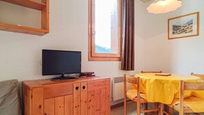 Une pièce comprenant un meuble en bois, un téléviseur, une table à manger recouverte d’une nappe jaune et une fenêtre avec des rideaux.