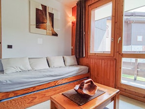 A room with a wooden floor, a bed with white bedding, a wooden table with a decorative object, a window with curtains, and a wall-mounted light fixture.