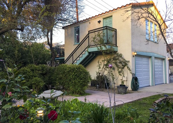 Una casa de dos pisos con un balcón verde, un garaje y un jardín con diversas plantas y flores.