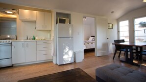 Eine kompakte Küche mit weißen Schränken, einem Kühlschrank und einem Herd. Es gibt eine Essecke mit Tisch und Stühlen, und im angrenzenden Raum steht ein Bett.