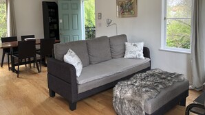 Ein modernes Wohnzimmer mit grauem Sofa, einem flauschigen Ottomanen, einem Essbereich mit Holztisch und -stühlen sowie einem großen Fenster mit Blick auf Grünflächen.