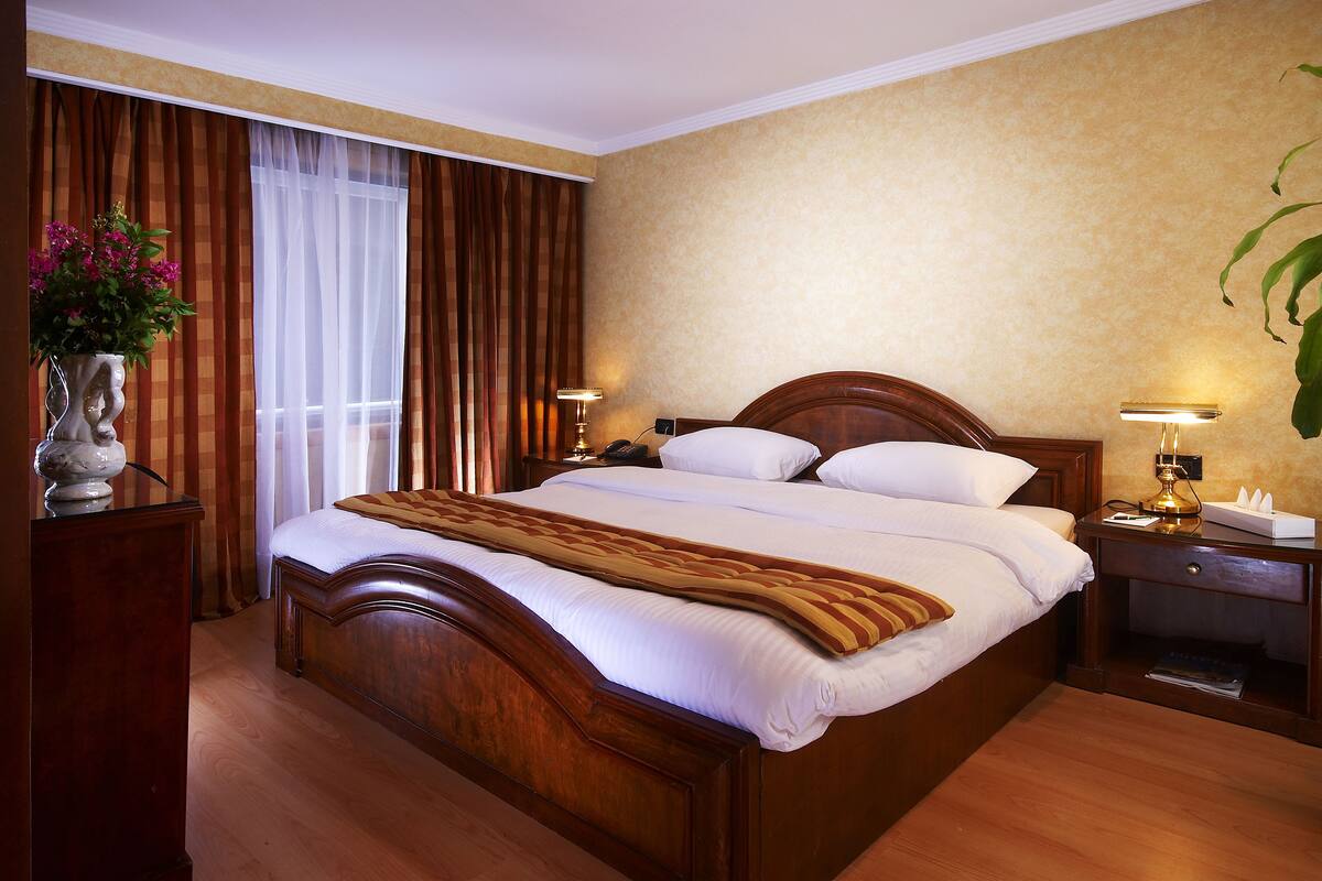 A hotel room with a large bed, wooden headboard, two bedside tables with lamps, a vase with flowers, and a window with curtains.