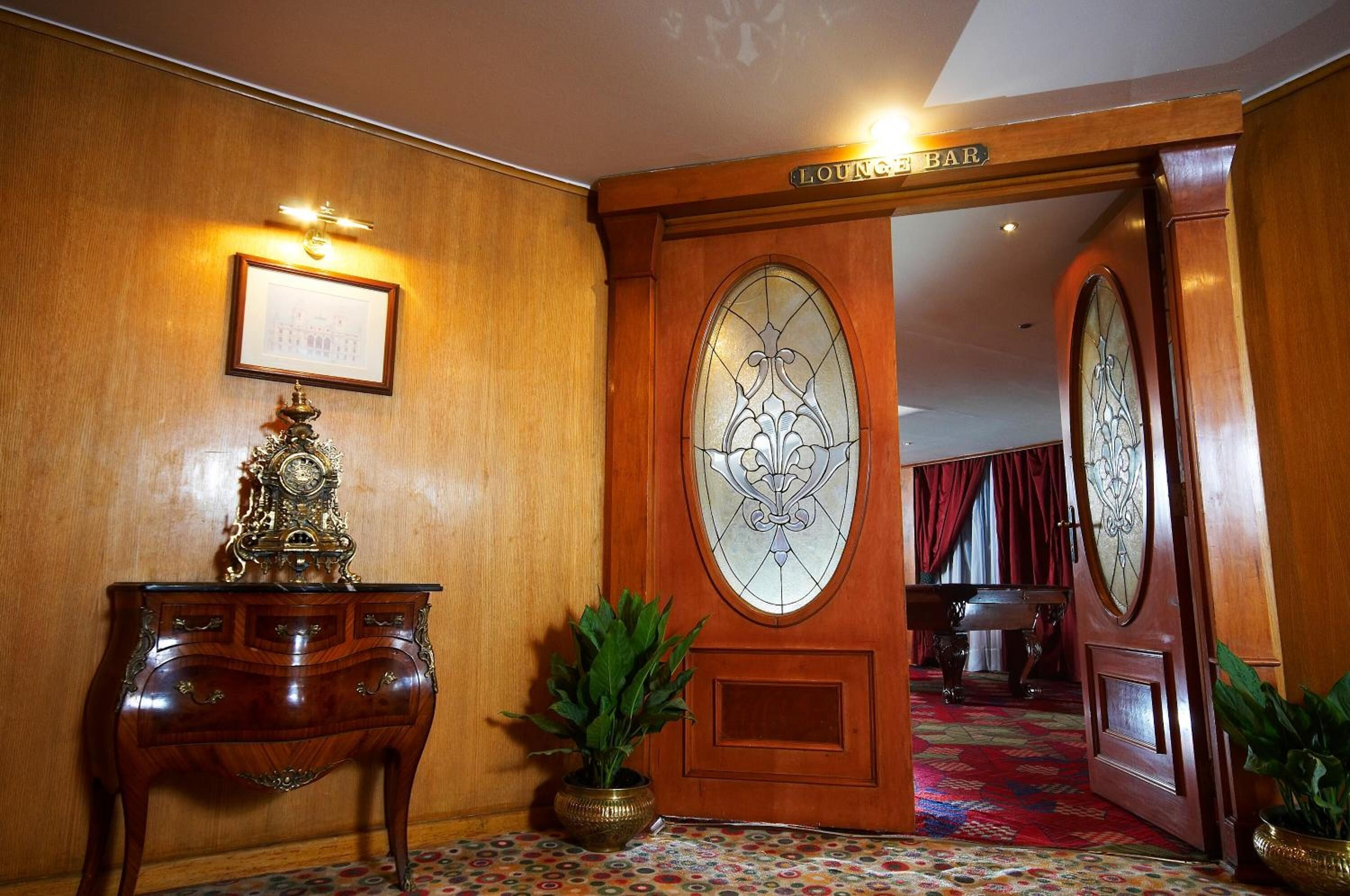 A wooden door with a sign reading "Lounge Bar," a framed picture on the wall, a decorative clock, and a patterned carpet.
