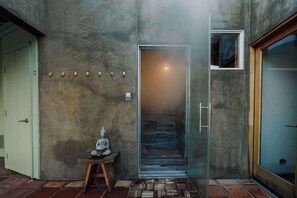 Un intérieur moderne avec un mur en béton, une petite statue de Bouddha posée sur une table en bois et une porte en verre donnant sur un couloir.