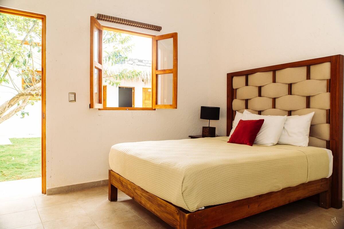 Un dormitorio con cama de madera, mesita de noche, lámpara y ventana con persiana abierta.
