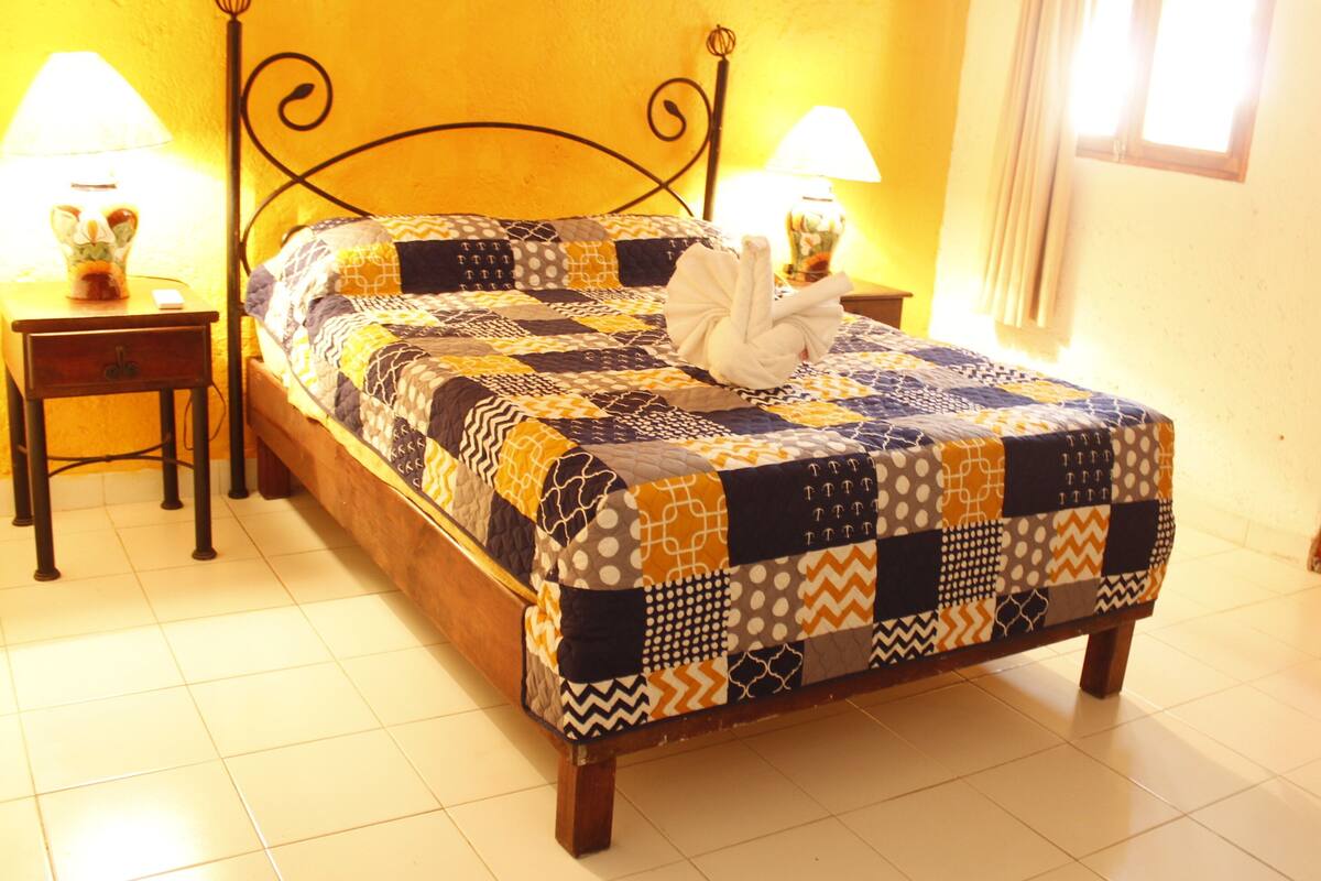 Una cama con colcha estampada, dos mesitas de noche con lámparas y una puerta de madera en una habitación con paredes amarillas y suelo de baldosas.