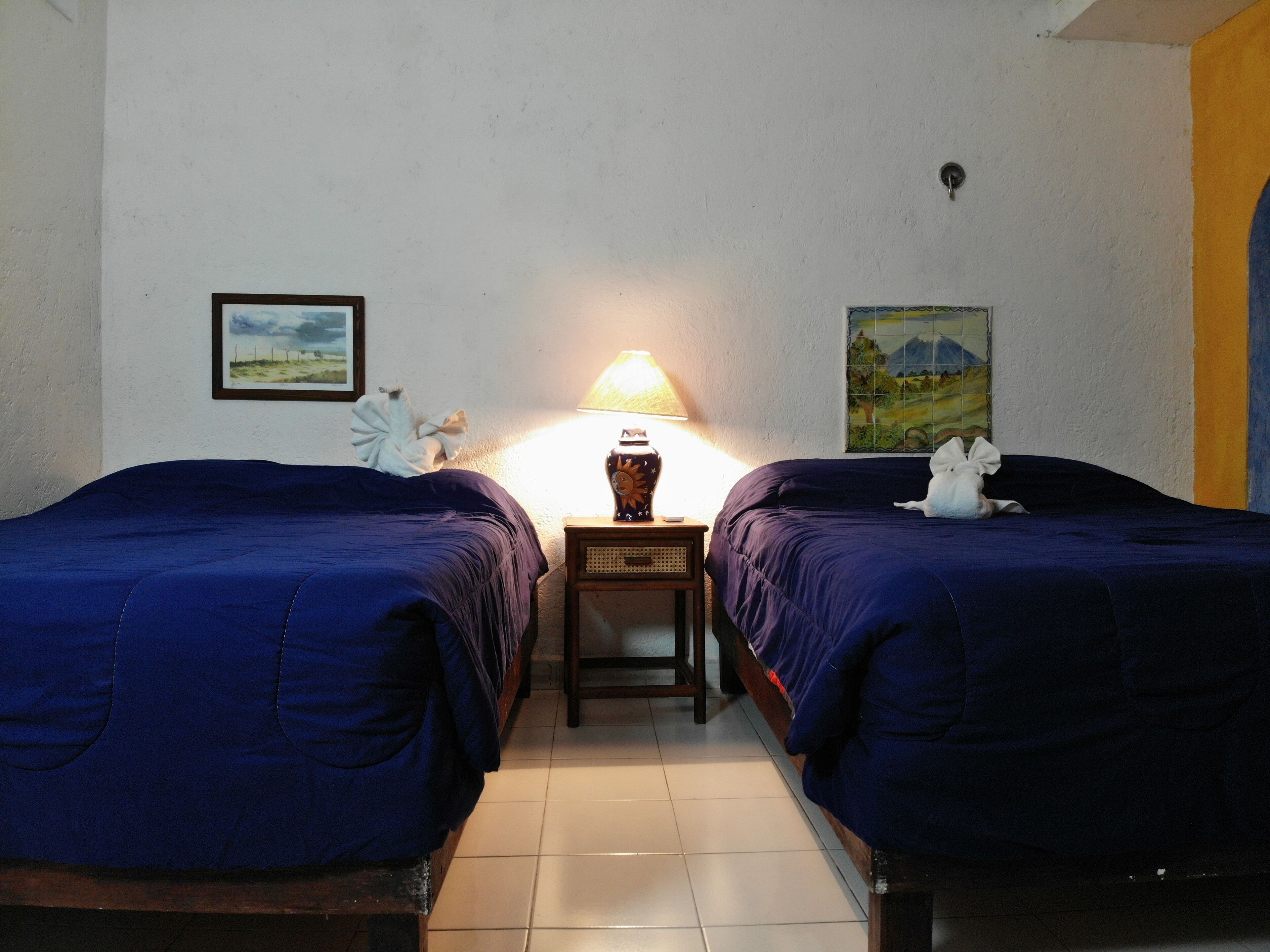 A room with two beds, a nightstand with a lamp, a framed picture on the wall, and a tiled floor.