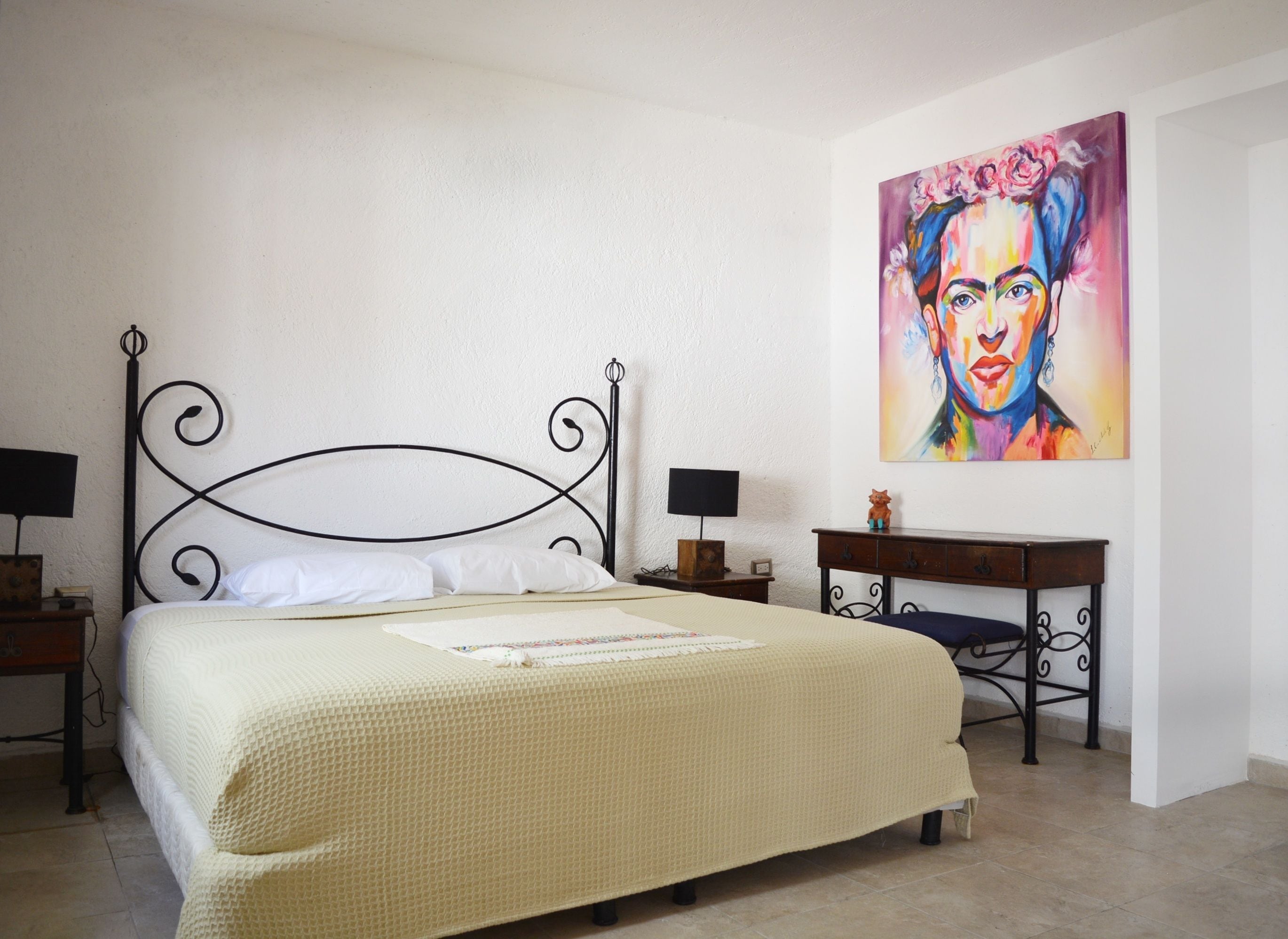A bed with a white quilt, a nightstand with a lamp, a desk with a chair, and a painting on the wall.