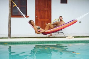 Two people relaxing on a hammock by a pool with a wooden door and window in the background.