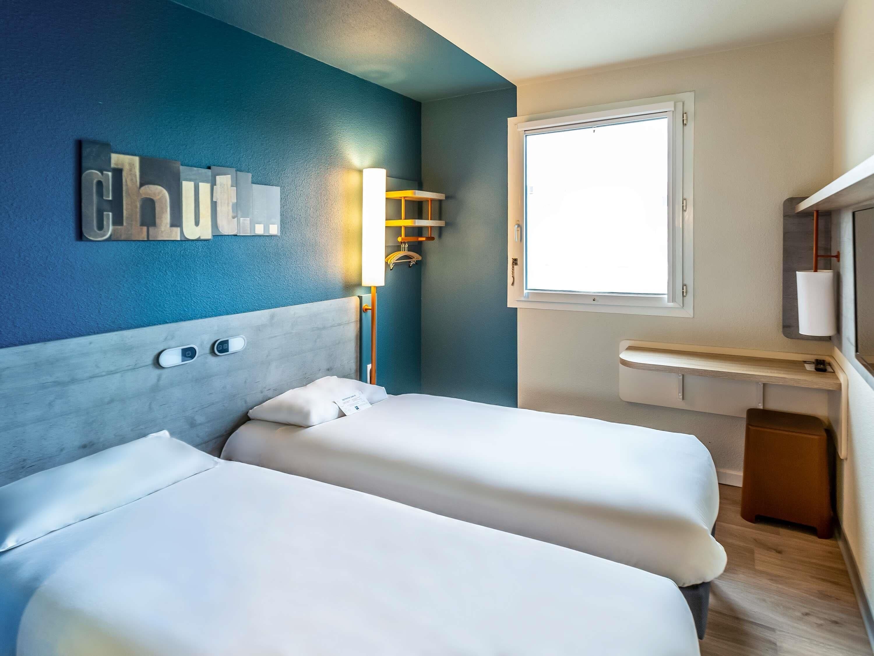 A hotel room with two beds, a nightstand, a window, and a wall-mounted light fixture.