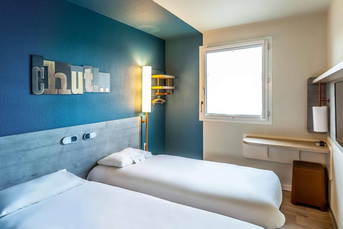 A hotel room with two beds, a nightstand, a window, and a wall-mounted light fixture.
