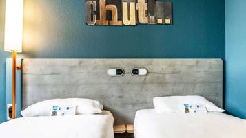 Habitación de hotel con pared azul, cabecero gris, dos camas y un letrero en la pared que dice 'chut...'.