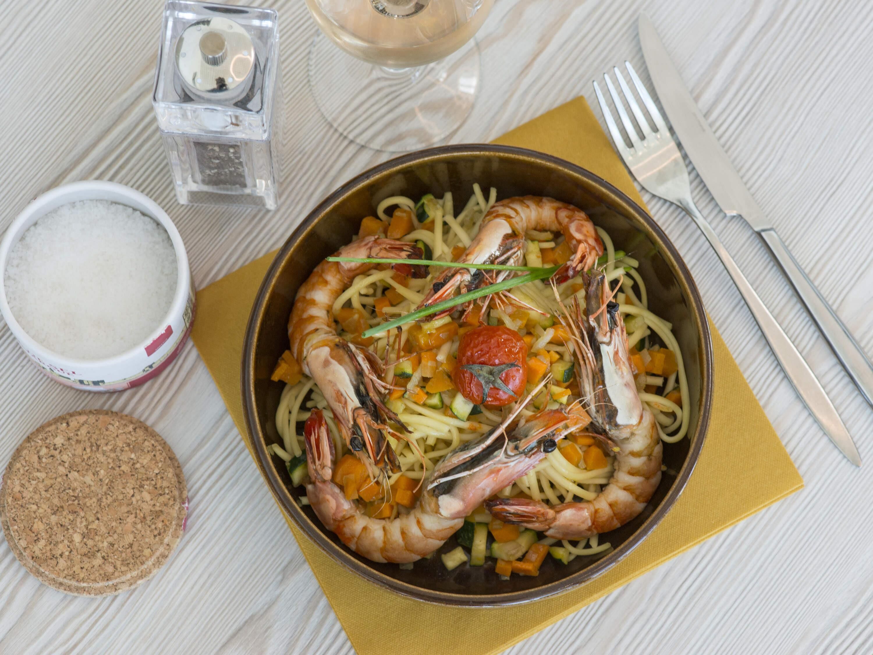 A bowl of pasta with shrimp, a salt shaker, a glass of water, and a cork coaster on a table.