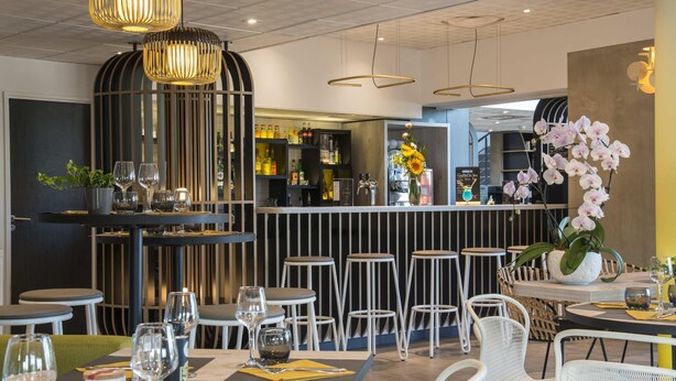 A restaurant interior with tables, chairs, hanging pendant lights, a bar area, and decorative plants. - ibis budget Pertuis Portes du Luberon (Pertuis)