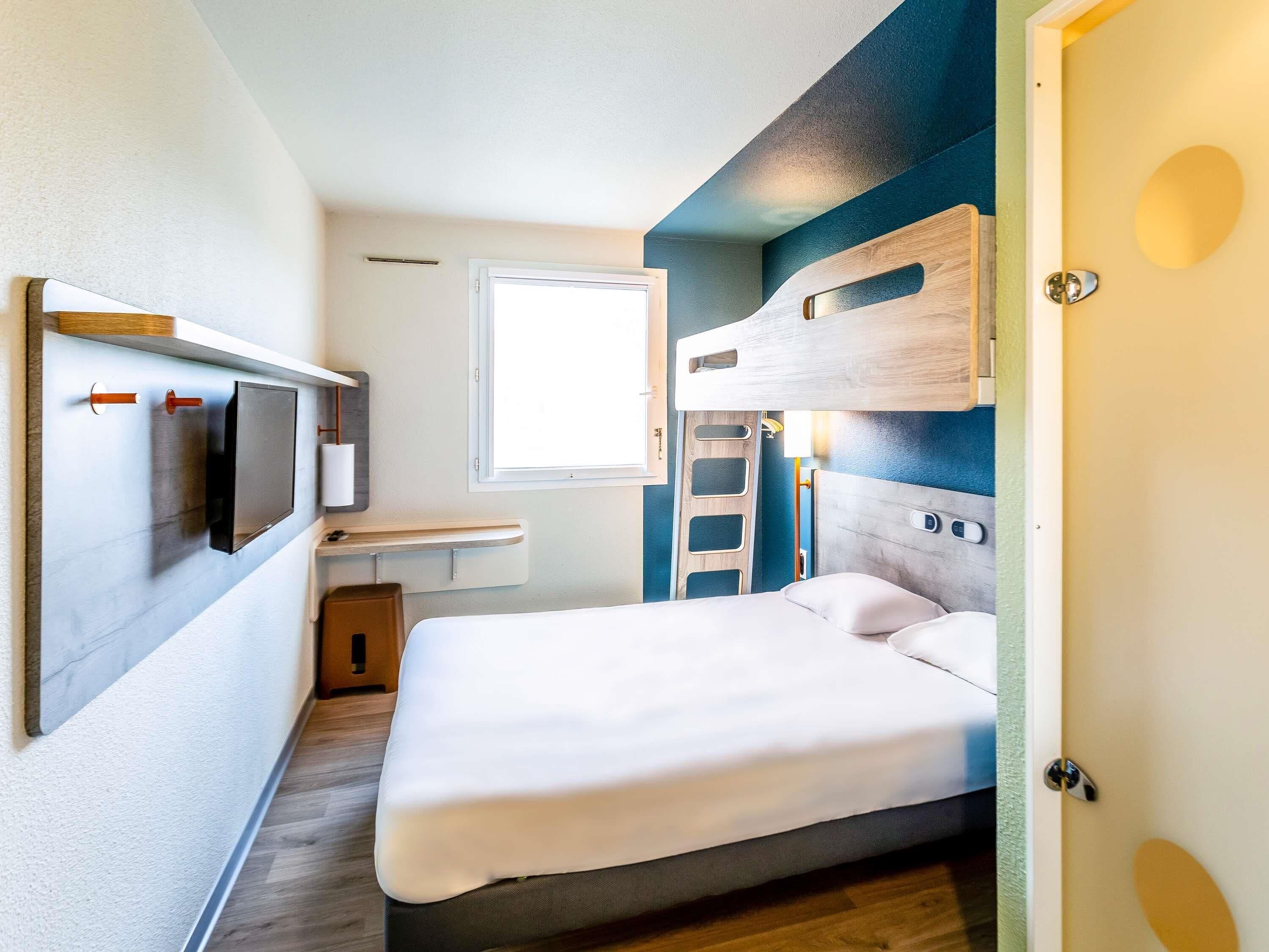 A compact room with a bed, a small table, a wall-mounted TV, and a window.