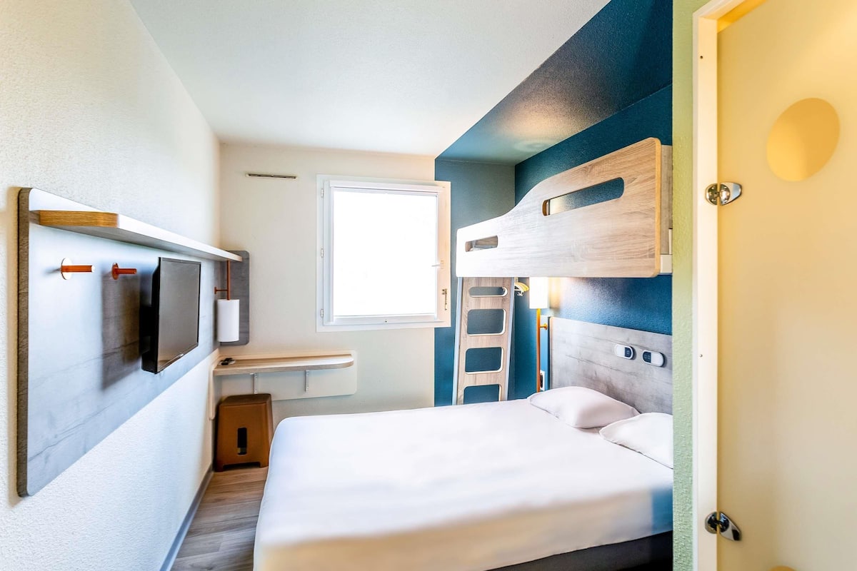 A compact room with a bed, a small table, a wall-mounted TV, and a window.