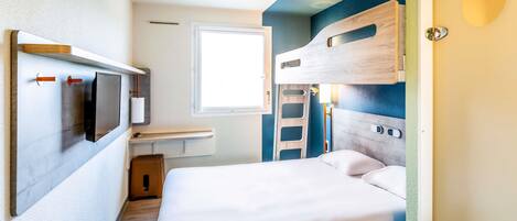 Una habitación pequeña con cama, una mesita, televisor en la pared y ventana.