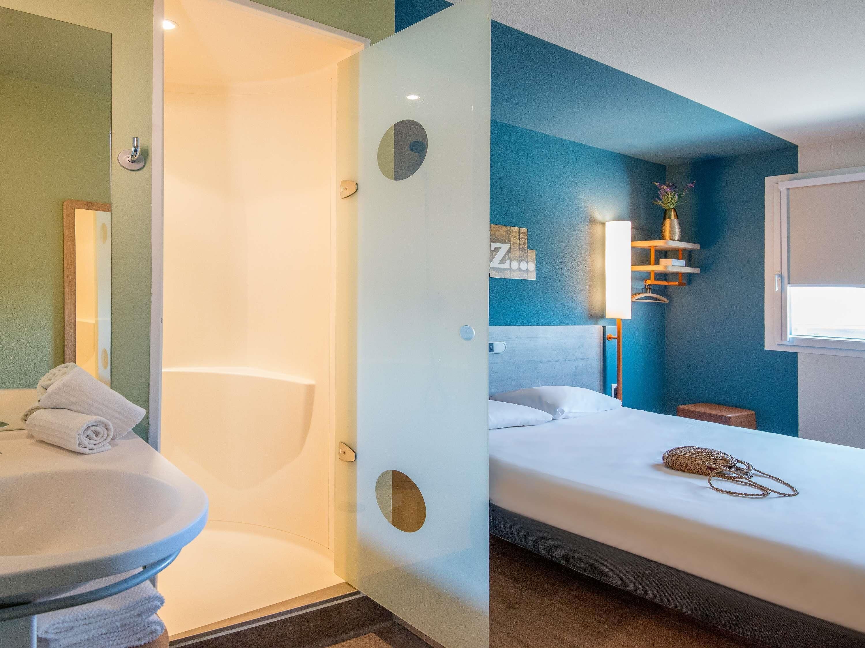 Una habitación de hotel moderna con baño, una cama con cabecera, una mesita de noche y un ventanal.